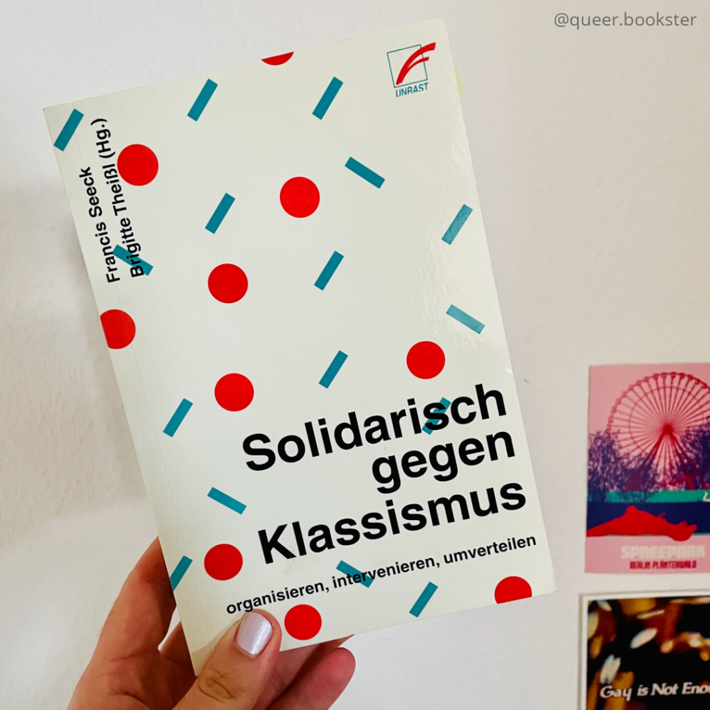 Eine Hand hält das Buchcover von Francis Seeck/ Brigitte Theißl »Solidarisch gegen Klassismus« vor einer Wand mit Postkarten.