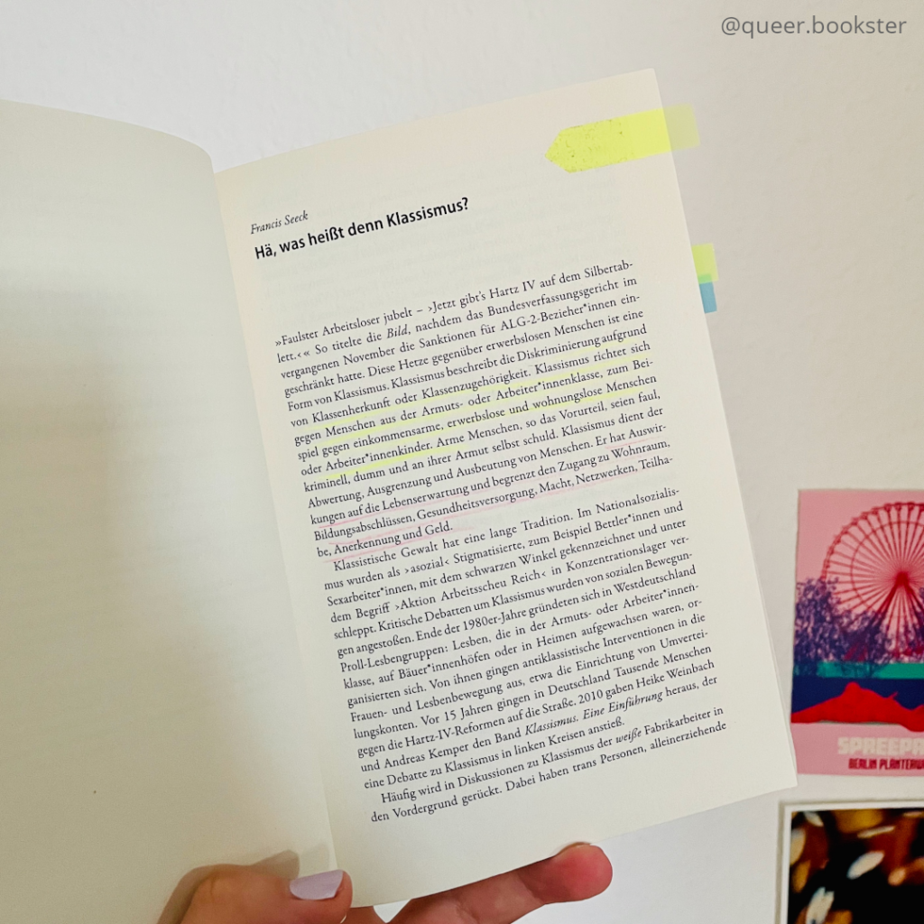 Eine Hand hält die aufgeschlagene Innenseite von Francis Seeck/ Brigitte Theißl »Solidarisch gegen Klassismus« vor einer Wand mit Postkarten.